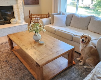 May include: A living room scene featuring a large, rectangular wooden coffee table with a lower shelf. The table is positioned on a patterned rug, with a beige sofa and a golden retriever dog. A fireplace and artwork are visible in the background.