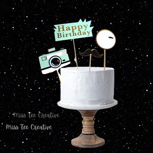 Puede incluir: Un pastel blanco con una cámara, un bigote y un adorno de "Feliz cumpleaños". El pastel está sobre un soporte para pasteles de madera sobre un fondo negro con estrellas blancas.