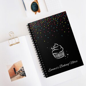 May include: Black spiral notebook with a white cupcake design and the text "Emm's Baking Notes". The cover is decorated with colourful sprinkles.