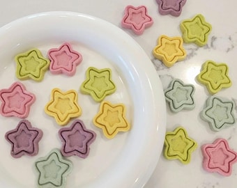 May include: A white plate with a variety of star-shaped cookies in different colors, including pink, yellow, green, and purple. The cookies are arranged in a circle on the plate. There are also some cookies on the table next to the plate.