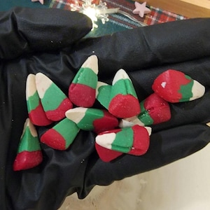 May include: A handful of colorful, tri-colored candies, resembling miniature Christmas trees. Each candy features layers of red, green, and white, with a pointed top. The candies are held in a black gloved hand, suggesting a festive theme.