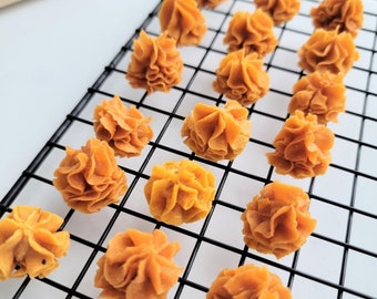 Puede incluir: Primer plano de una rejilla de alambre con 17 pequeños pasteles redondos y rizados de color naranja. Los pasteles están dispuestos en filas en la rejilla.