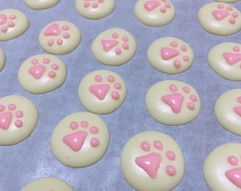 May include: Overhead shot of numerous round, pale yellow macarons, each decorated with a pink paw print design. The macarons are arranged on a light-colored surface, creating a visually appealing pattern. The pink paw prints add a playful touch.