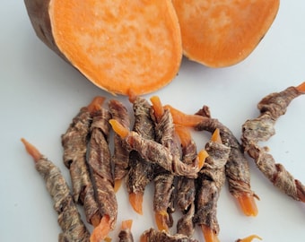 Peut inclure: Gros plan sur un tas de friandises pour chien à la patate douce séchée. Les friandises sont brunes et orange et ont une forme torsadée. Il y a deux moitiés de patate douce en arrière-plan.