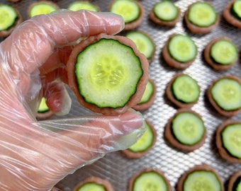 Peut inclure: Gros plan sur une main tenant une petite collation ronde de couleur rose surmontée d'une tranche de concombre. La collation est entourée d'autres collations similaires sur un plateau argenté.