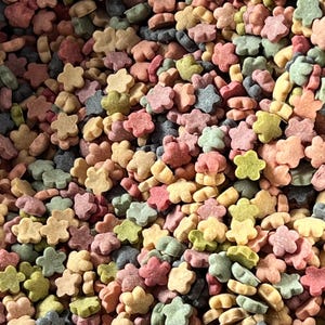 May include: A close-up shot of a pile of colorful, flower-shaped sprinkles. The sprinkles are in various pastel colors, including pink, yellow, green, and blue. They are scattered randomly, creating a textured and visually appealing pattern. These are likely used for decorating baked goods.