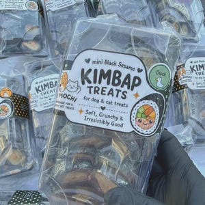 May include: A clear plastic package of mini Black Sesame Kimbap Treats for dog and cat treats. The label features a cartoon kimbap roll, a duck icon, and the text: mini Black Sesame Kimbap Treats for dog & cat treats, Soft, Crunchy & Irresistibly Good.
