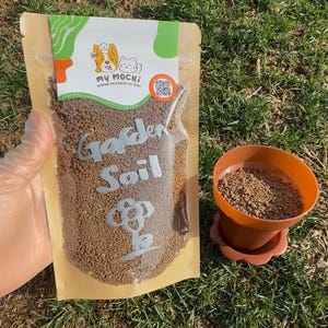 May include: A brown paper bag with a clear plastic window, filled with brown garden soil. The bag has a white label with the text "Garden Soil" and a logo for "My Mochi" with a dog and cat wearing chef hats. A small brown plastic pot with a saucer is filled with the same soil.