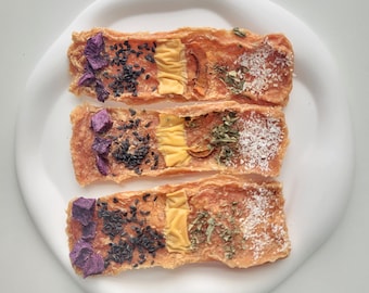 Peut inclure: Trois friandises rectangulaires pour chien avec différentes garnitures. Les friandises sont à base de viande séchée et ont une couleur brun clair. Les garnitures comprennent des fruits secs, des herbes et des graines de sésame noir.