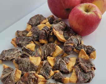 Peut inclure: Un tas de tranches de pommes séchées, dont certaines sont roulées, sur une surface blanche. Trois pommes rouges sont en arrière-plan.