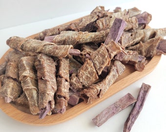 Peut inclure: Un bol en bois rempli de friandises pour chiens à base de viande séchée et de patate douce violette. Les friandises sont disposées en tas et certaines sont éparpillées sur une surface blanche.