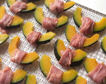 Peut inclure: Gros plan sur une grille métallique avec des tranches de citrouille enveloppées de fines tranches de viande. Les tranches de citrouille sont jaunes et vertes, et la viande est rose.