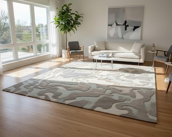 May include: A large, rectangular area rug with a unique, abstract pattern in shades of beige, gray, and light blue dominates the hardwood floor of a living room. The room features a white sofa, chairs, and a round coffee table, creating a modern, minimalist aesthetic.