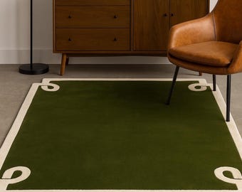 Op de afbeelding: Een rechthoekig groen tapijt met een crèmekleurige rand en decoratieve hoekaccenten. Het tapijt ligt op een betonnen vloer, met een houten kast en leren stoel op de achtergrond. Afmetingen: ongeveer 152 x 229 cm.