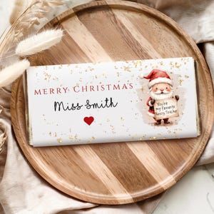 May include: A festive chocolate bar with "Merry Christmas Miss Smith" in red and black text, a red heart, and gold glitter. A Santa Claus illustration holds a sign saying "You're my favourite Nursery Teacher".