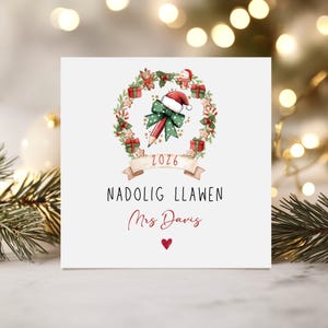 May include: A square white greeting card featuring a Christmas wreath illustration with a pencil wearing a Santa hat, red gift boxes, and gingerbread cookies. Below the wreath is a banner with 2026, the text NADOLIG LLAWEN, Mrs Davis, and a red heart.