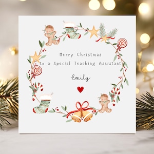 May include: A square white Christmas greeting card featuring a central wreath made of watercolour illustrations including gingerbread people, stockings, candy canes, lollipops, stars, and bells. The text reads Merry Christmas to a Special Teaching Assistant Emily with a red heart.