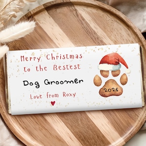 May include: A rectangular chocolate bar wrapper with a white background and gold speckles. It features the red text Merry Christmas to the Bestest Dog Groomer, Love from Roxy, a red heart, and a brown dog paw print wearing a red Santa hat with 2026.