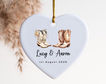 May include: A white heart-shaped ceramic ornament hanging from a gold string. The design features two pairs of brown cowboy boots, one adorned with floral accents. The text reads "Lucy & Aaron" in script and "1st August 2026" in a sans-serif font.