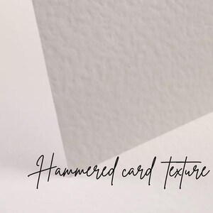 May include: Close-up of a textured, off-white paper surface with the words "Hammered card texture" in cursive script. The paper has a subtle, uneven surface, and the lighting creates soft shadows.