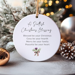 May include: A round white ceramic Christmas ornament hanging from a gold ribbon. The text reads A Scottish Christmas Blessing, Blessed be your Christmas, Cosy be your hearth, Merry be your family, Peaceful be your heart, with a small purple thistle graphic.