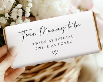 May include: A white rectangular gift with the text "Twin Mummy to be" in elegant script. Below, it reads "Twice as special, twice as loved" with a heart symbol. The gift is held in a hand, with a floral background.