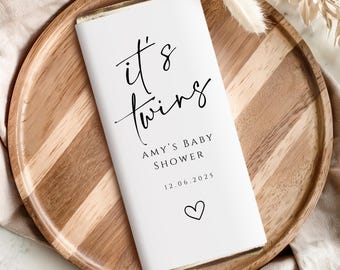 May include: A white invitation with the text "it's twins" in black script, along with "AMY'S BABY SHOWER" and the date 12.06.2025. A heart symbol is at the bottom. The invitation rests on a wooden tray.