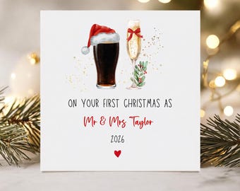May include: A white greeting card featuring a dark beer in a glass with a Santa hat, a champagne flute with a red bow and holly, and the text: ON YOUR FIRST CHRISTMAS AS Mr & Mrs Taylor 2026 with a small red heart at the bottom center.