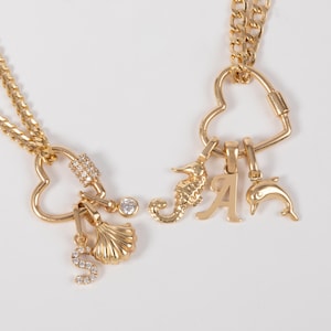 May include: Two gold-toned necklaces featuring heart-shaped carabiner clasps. One necklace includes a crystal-encrusted letter S, a seashell charm, and a small round crystal. The other features a seahorse, the letter A, and a dolphin charm.