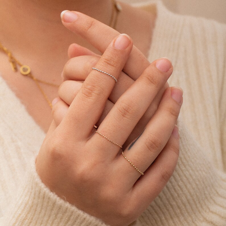 K&ouml;nnte beinhalten: Eine Hand tr&auml;gt drei zarte goldene und silberne Perlenringe. Die Ringe sind d&uuml;nn und schlicht, mit kleinen Perlen, die einen dezenten Glanz erzeugen.