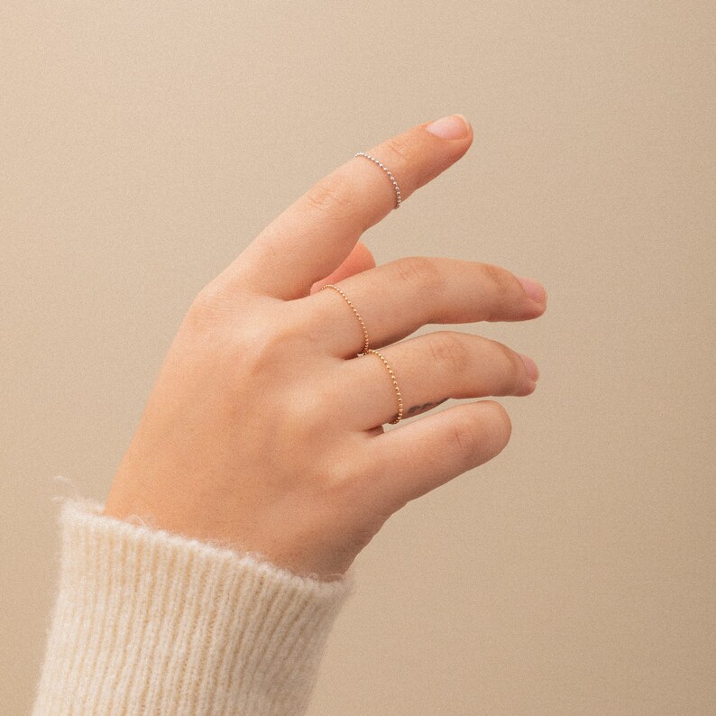 K&ouml;nnte beinhalten: Eine Hand tr&auml;gt drei zarte Ringe aus Gold und Silber. Die Ringe sind d&uuml;nn und haben ein perlenf&ouml;rmiges Design.