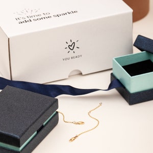 Pode incluir: Um colar de ouro com uma corrente e fecho delicados &eacute; exibido perto de duas caixas de joias abertas. As caixas s&atilde;o azul marinho com interior azul claro. Uma caixa branca com o texto "It's time to add some sparkle" est&aacute; no fundo.