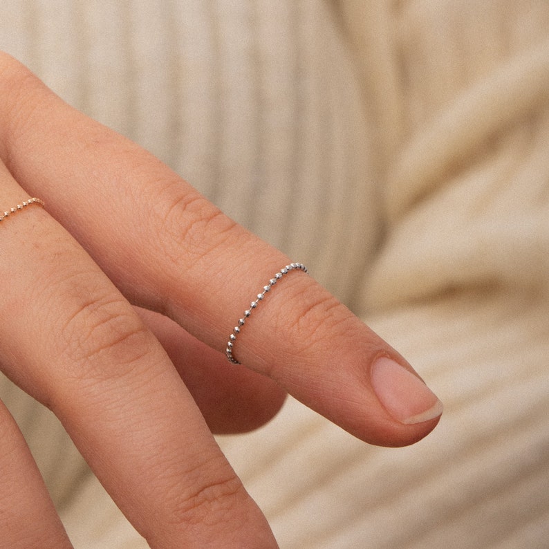 K&ouml;nnte beinhalten: Ein zarter Silberring mit einem Perlenketten-Design. Der Ring wird am Finger einer Hand getragen.