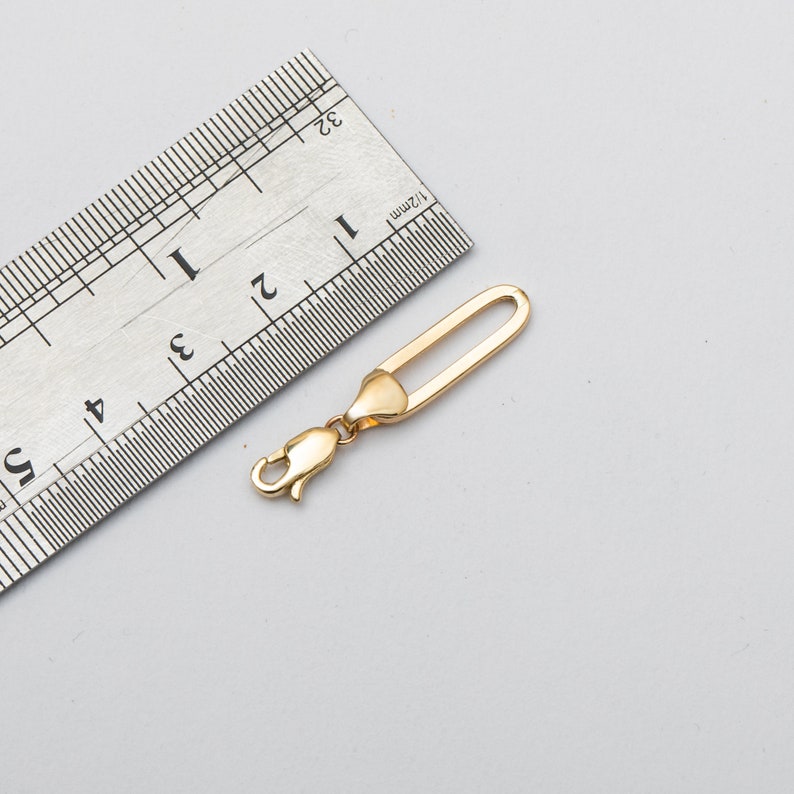 Pode incluir: Um fecho de joia dourado, com aproximadamente 3,8 cm de comprimento, com um fecho de lagosta numa extremidade e um la&ccedil;o arredondado e alongado na outra. O fecho &eacute; mostrado ao lado de uma r&eacute;gua.
