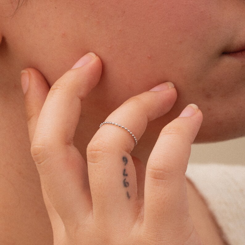 K&ouml;nnte beinhalten: Nahaufnahme einer Hand, die einen d&uuml;nnen silbernen Perlenring am Ringfinger tr&auml;gt. Der Ring ist schlicht und zart, mit kleinen, gleichm&auml;&szlig;ig verteilten Perlen.