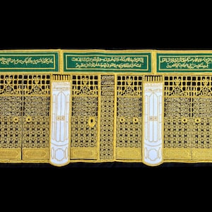 May include: A close-up of a black and gold embroidered fabric with Arabic text. The fabric is divided into three sections, each with a different design. The fabric is likely a religious artifact.
