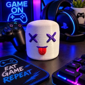 May include: A white cylindrical object with a 3D-printed texture featuring a purple X-shaped eye design and a purple mouth with a protruding red tongue. The object is positioned in a gaming environment with a controller, headset, and keyboard.