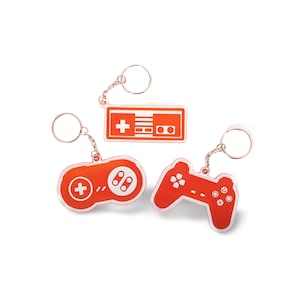 Puede incluir: Tres llaveros naranjas con diseños blancos. Los llaveros tienen forma de mando de videojuego, consola de videojuego clásica y mando de videojuego moderno.
