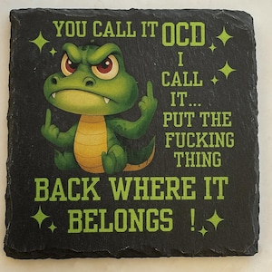 May include: Black square slate coaster with green text and a cartoon alligator giving the middle finger. The text says "YOU CALL IT OCD, I CALL IT... PUT THE FUCKING THING BACK WHERE IT BELONGS!"