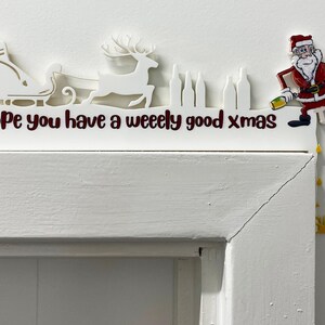 May include: White Christmas decoration with a Santa Claus figure holding a beer, a reindeer pulling a sleigh, and the text "Hope you have a weeelly good xmas".