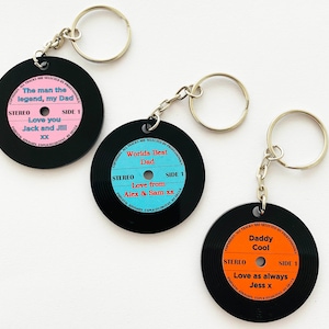May include: Three black vinyl record-shaped keychains with colorful labels. The first keychain says "The man the legend, my Dad" with pink and orange text. The second keychain says "Worlds Best Dad" with blue and orange text. The third keychain says "Daddy Cool" with orange text.