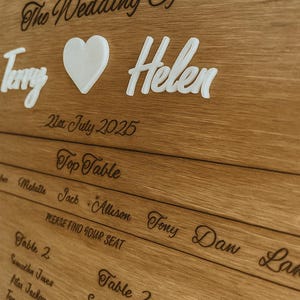 May include: A wooden wedding seating chart with the names "Terry" and "Helen" in white 3D lettering, a white heart, and the date "21st July 2025". The chart includes table assignments and guest names in elegant script.