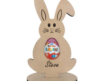 Puede incluir: Porta huevos de Pascua de madera en forma de conejo con un huevo de chocolate Kinder Surprise en su interior. El soporte tiene una base y el nombre "Steve" está escrito en la parte delantera.