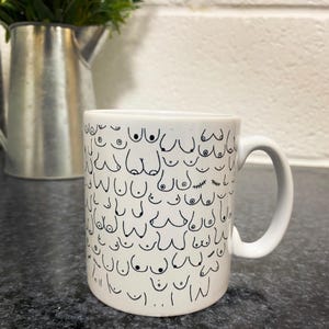 May include: White ceramic mug with a black and white pattern of stylized breasts. The mug has a handle on the right side.