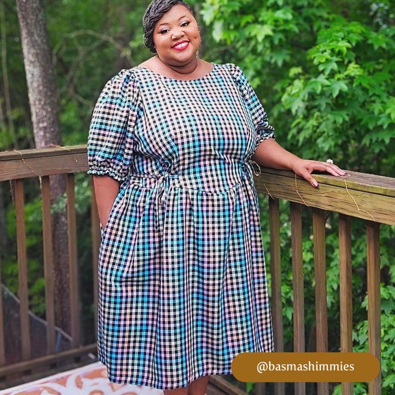 May include: A woman wearing a black and white gingham dress with a blue and pink plaid pattern. The dress has a cinched waist and puffed sleeves. The woman is standing on a wooden deck and smiling at the camera. The text "@basmashimmiies" is visible in the lower right corner of the image.