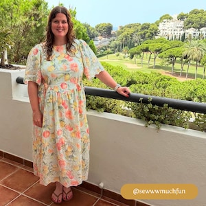 May include: A woman wearing a light blue dress with a white and yellow floral pattern. The dress has a ruffled neckline and short sleeves. She is standing on a balcony overlooking a golf course.