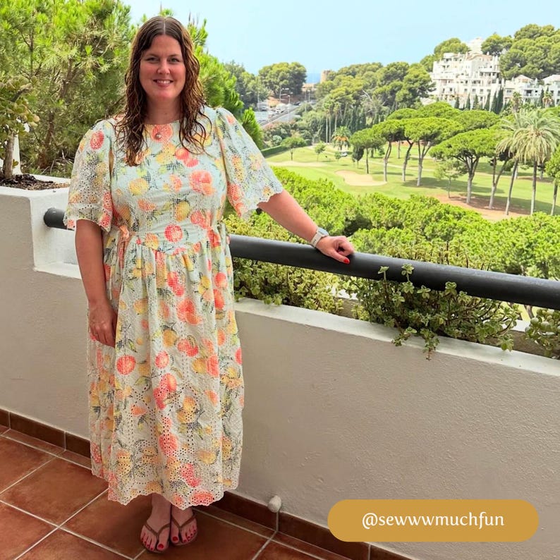 May include: A woman wearing a light blue dress with a white and yellow floral pattern. The dress has a ruffled neckline and short sleeves. She is standing on a balcony overlooking a golf course.
