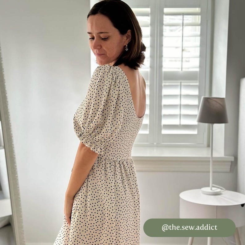 May include: A white dress with black polka dots and a scoop neckline. The dress has puffed sleeves and a fitted bodice. The dress is shown on a model in front of a window with white blinds. The text "@the.sew.addict" is visible in the lower right corner of the image.