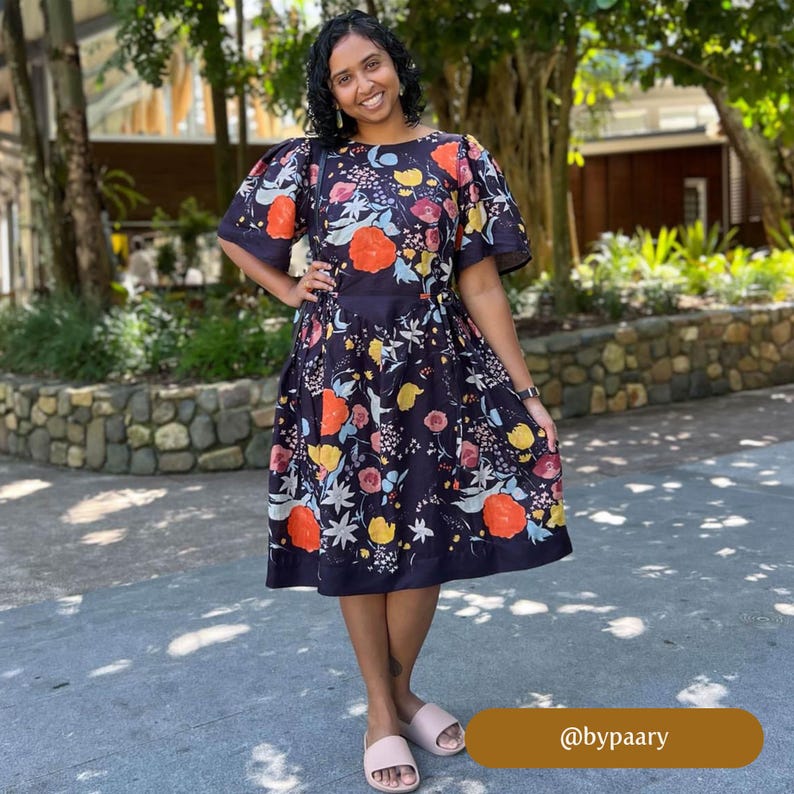 May include: A woman wearing a black midi dress with a floral print. The dress has a round neckline and short sleeves. The woman is standing in front of a stone wall and has her hair pulled back. The caption at the bottom of the image reads "@bypaary".