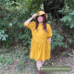 May include: A woman wearing a mustard yellow dress with a cinched waist and puff sleeves. She is wearing a straw hat with a sunflower on it and brown boots. The woman is standing in a wooded area. The text "@honeysucklehillhomestead" is at the bottom of the image.